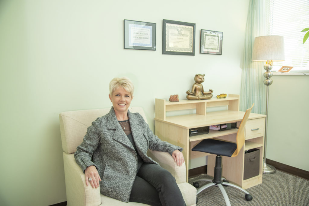 A woman in short haircut and grey long coat in her office, surrounded by certificates indicating their expertise